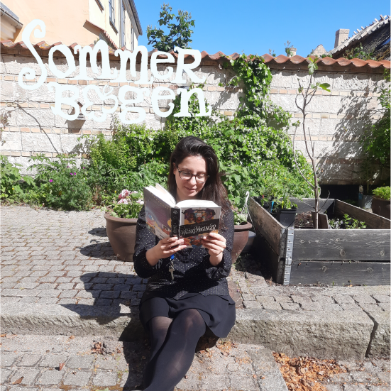 Sommerbogen. Børnebibliotekaren læser en bog i biblioteksgården.