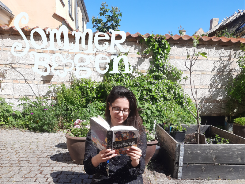 Sommerbogen. Børnebibliotekaren læser en bog i biblioteksgården.