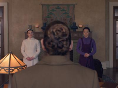 Scene i et rum: to kvinder står med foldede hænder, mens en person i forgrunden ses bagfra.